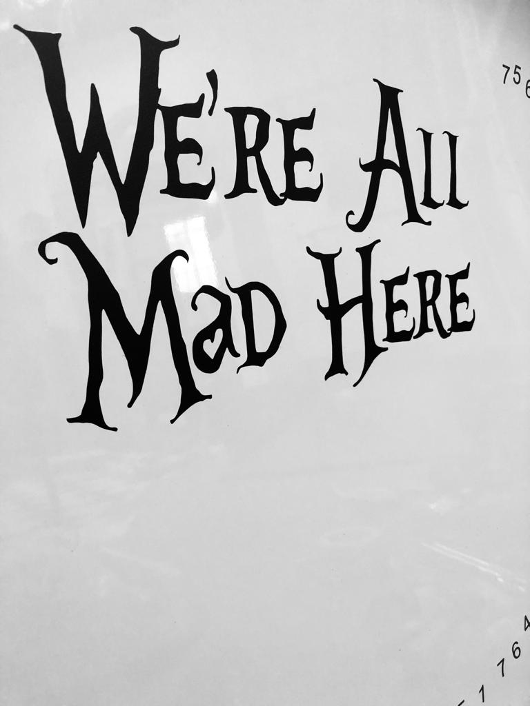 Black and white photo of a sign on the wall that says we're all mad here